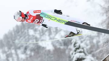 Skoczek narciarski w locie, w białym kombinezonie i z nartami Fischer, na tle zaśnieżonych drzew.