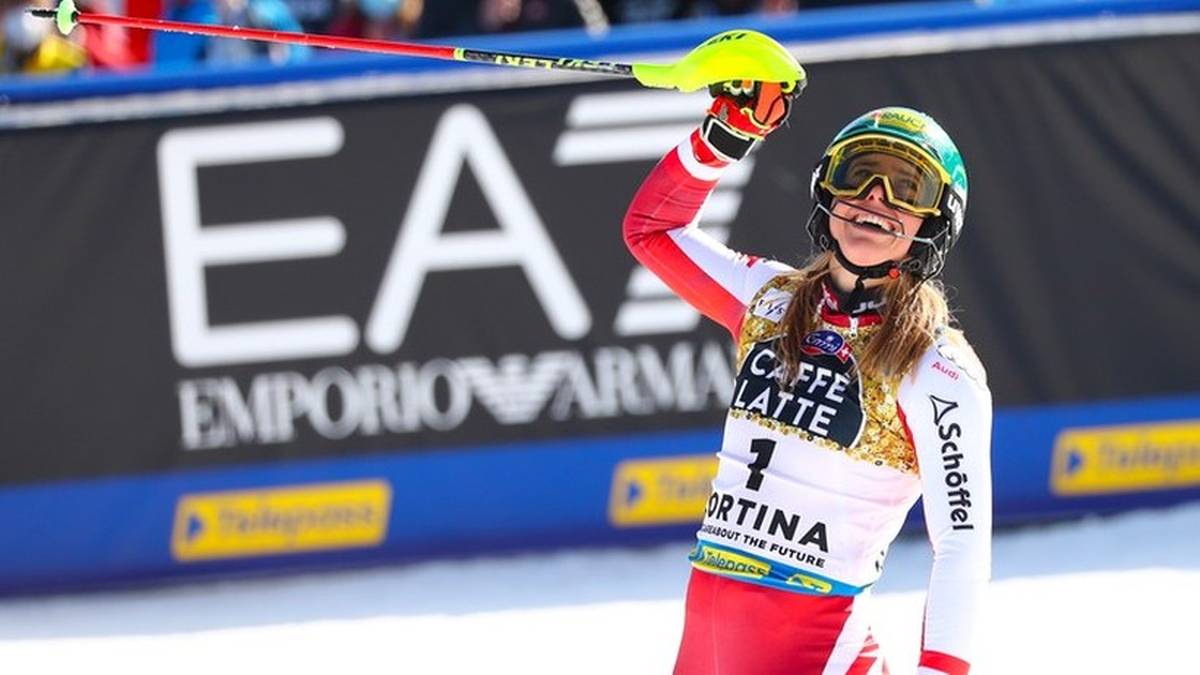 Narciarka alpejska w stroju sportowym z numerem startowym 1 na piersi, trzymająca kijki narciarskie i unosząca rękę w geście triumfu.