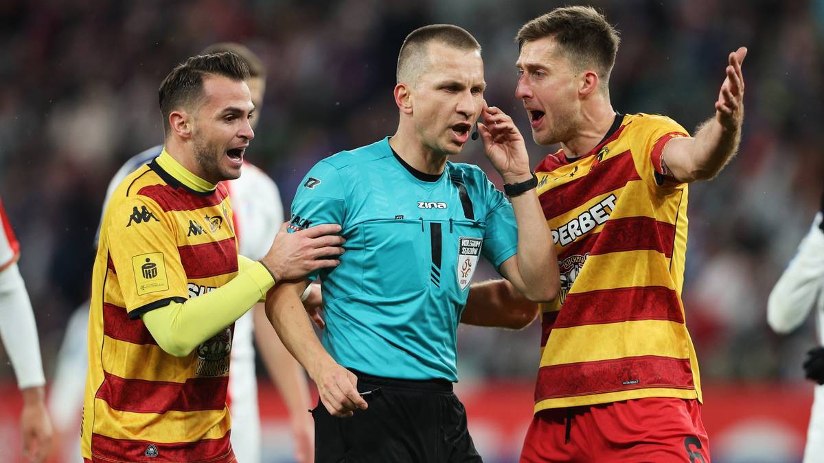 Sędzia w niebieskiej koszulce rozmawia przez telefon, otoczony przez dwóch piłkarzy w żółto-czerwonych strojach, którzy wydają się być zdenerwowani.