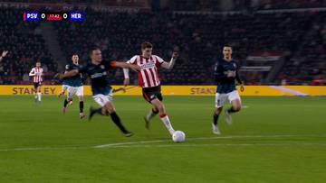 Piłkarz w czerwono-białej koszulce prowadzi piłkę na boisku, ścigany przez dwóch przeciwników w niebieskich strojach.