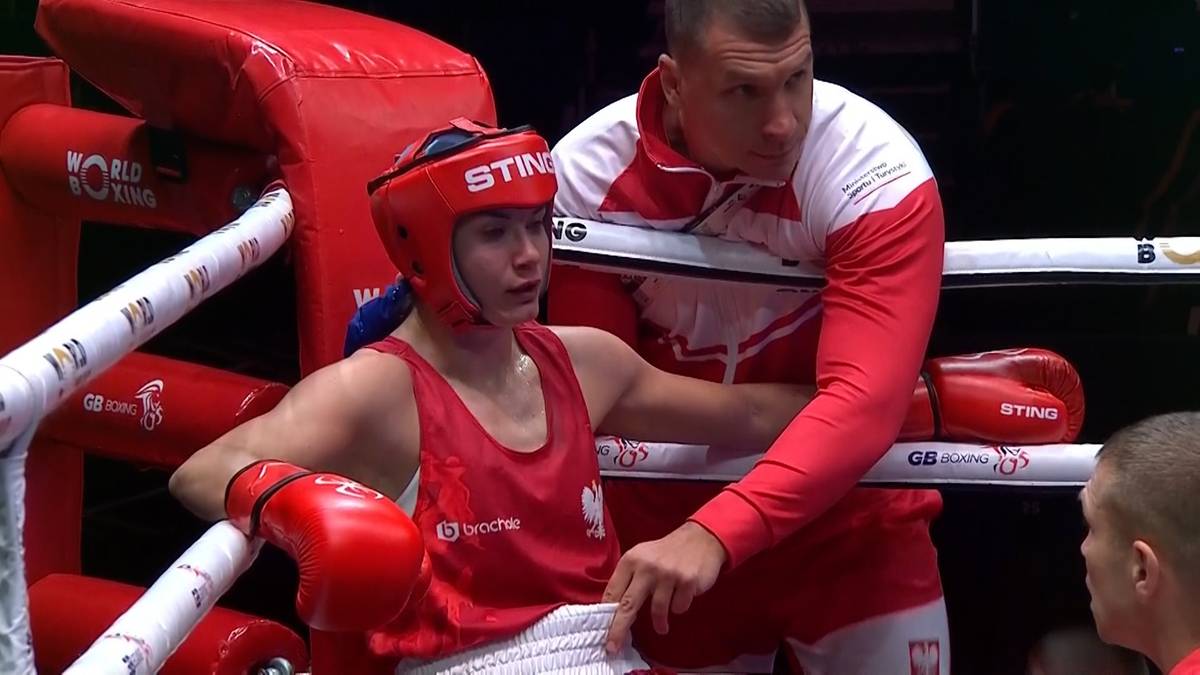 Młoda bokserka w czerwonym kasku i stroju, w rękawicach, stoi w ringu bokserskim. Obok niej stoi trener w czerwono-białej bluzie.