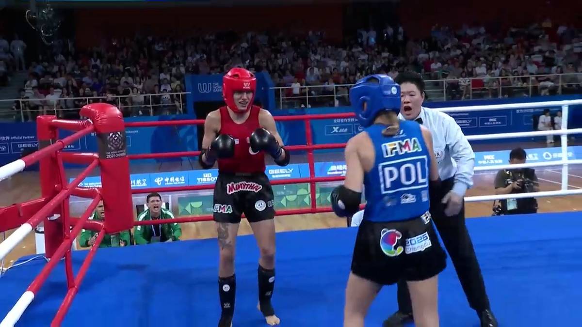 Dwie zawodniczki w niebieskich i czerwonych kaskach i rękawicach, rywalizują na ringu bokserskim. Jedna z nich, w niebieskim stroju z napisem "POL" na plecach, jest zwrócona tyłem do widza. Druga, w czerwonym stroju, stoi do niej bokiem. Obok nich znajduje się mężczyzna w białej koszuli, prawdopodobnie sędzia.