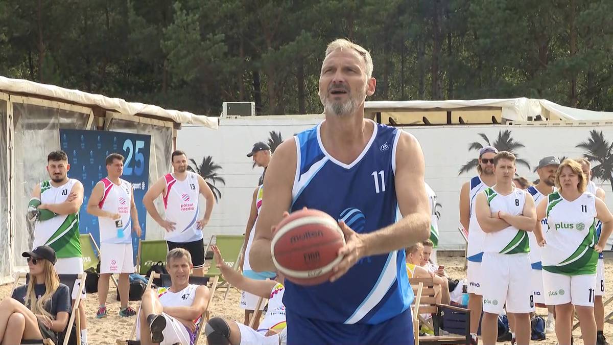 Mężczyzna w stroju sportowym trzyma piłkę do koszykówki, przygotowując się do gry. W tle widać innych graczy i publiczność na plaży.