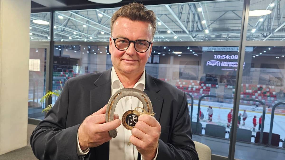 Mężczyzna w garniturze i okularach trzyma złoty medal w kształcie podkowy.