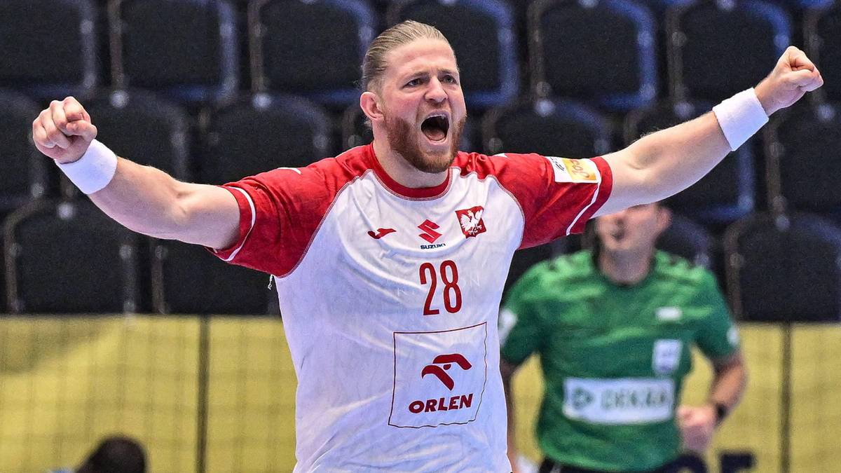 Piłkarz ręczny w białej koszulce z numerem 28 i czerwonymi rękawami, świętujący bramkę z uniesionymi rękami na parkiecie hali sportowej.