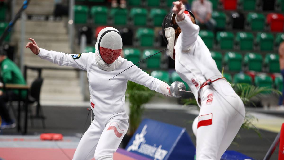 Dwie sportsmenki w białych strojach i maskach do szermierki toczą pojedynek na szpady. Jedna z nich, z biało-czerwoną flagą na rękawie, wykonuje wypad.