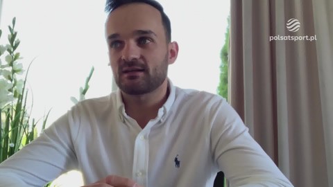 Bartosz Zmarzlik, polski żużlowiec, w białej koszuli z widocznym logo, siedzi przed kamerą. W tle zielona roślinność i zasłona z logo Polsat Sport.