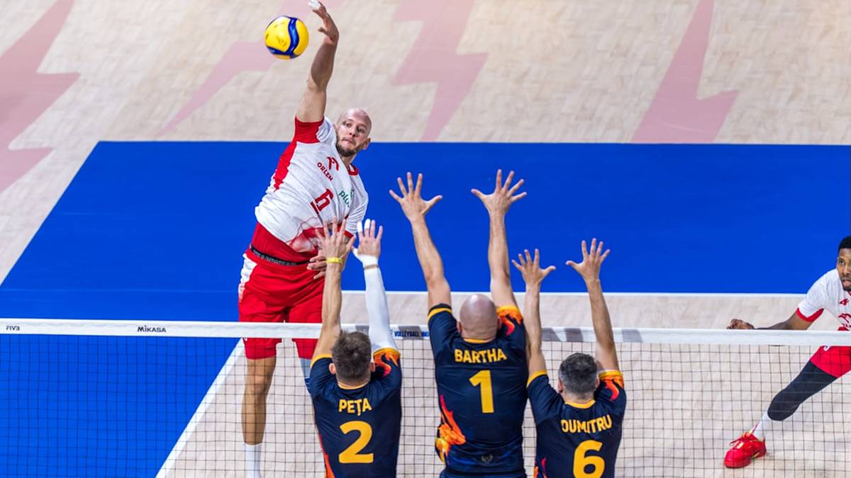 Siatkarz w białej koszulce z czerwonymi elementami i czerwonych spodenkach wyskakuje do ataku, uderzając piłkę ręką. Naprzeciwko niego widoczni są trzej zawodnicy drużyny przeciwnej z numerami na koszulkach: 2, 1 i 6, próbujący zablokować jego atak. Gra odbywa się na niebieskim boisku z białą siatką.