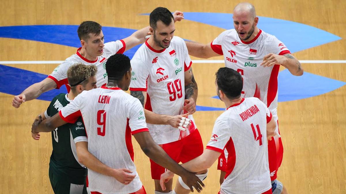 Sześciu graczy w biało-czerwonych strojach siatkarskich tworzy koło, patrząc na siebie i gestykulując. Dwóch z nich trzyma się za ręce. Jest to zdjęcie zrobione z góry podczas meczu.