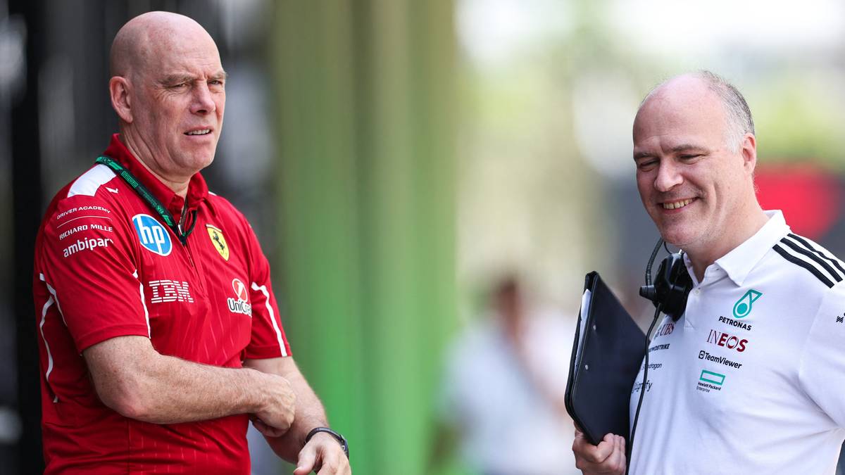 Dw&oacute;ch mężczyzn w strojach sportowych, jeden w czerwonym z logo Ferrari, drugi w białym z logo Mercedesa, rozmawia na tle toru wyścigowego.