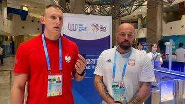 Dw&oacute;ch mężczyzn, jeden w czerwonej koszulce, drugi w białej, obie z przypinkami z godłem Polski, stoi przed tablicą informacyjną.