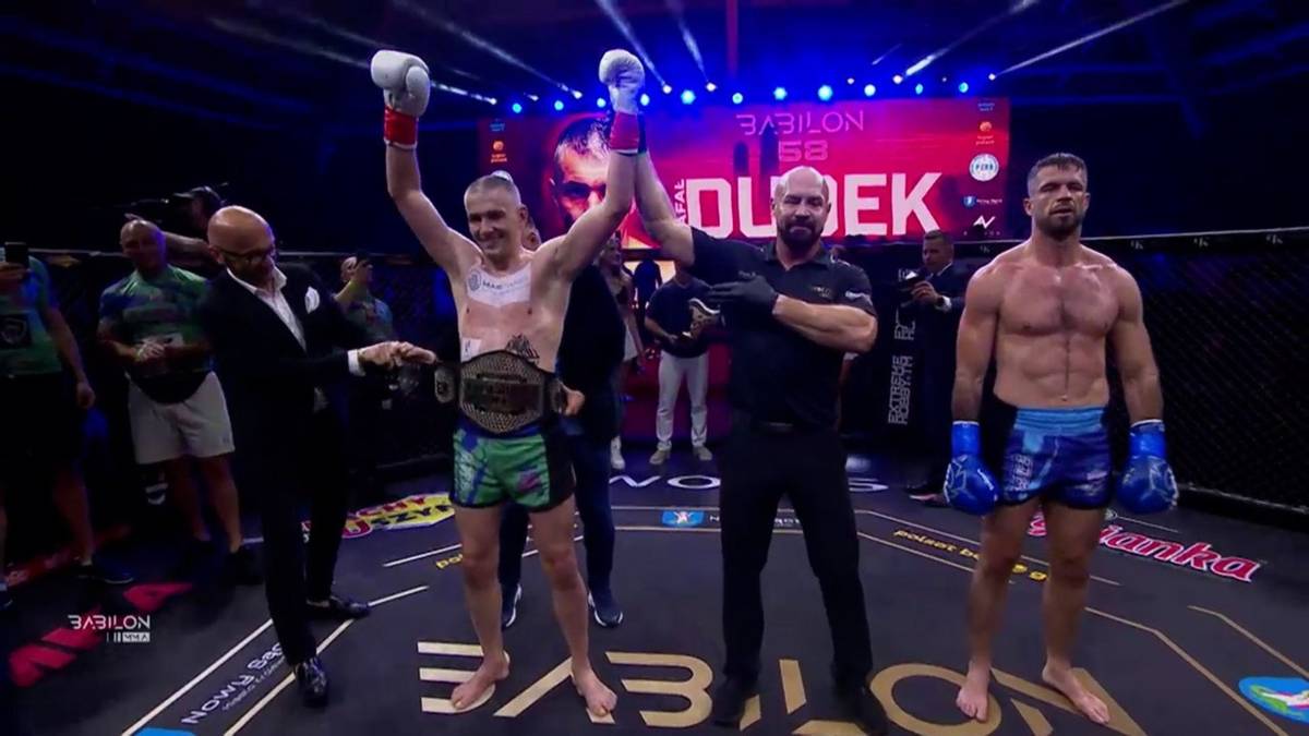 Dw&oacute;ch zawodnik&oacute;w MMA stoi w ringu, jeden z nich, Rafał Dudek, unosi ręce i prezentuje pas mistrzowski.