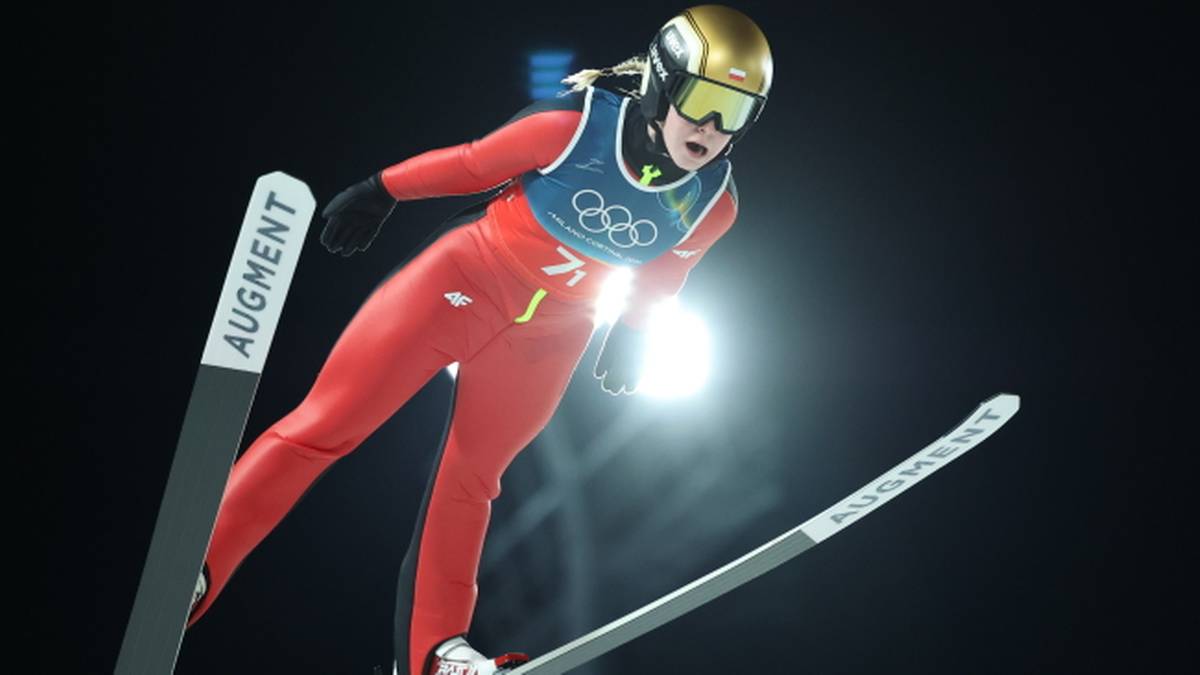 Skoczkini narciarska w czerwonym stroju wykonuje skok na nartach, ubrana w kask i gogle, w locie nad skocznią.