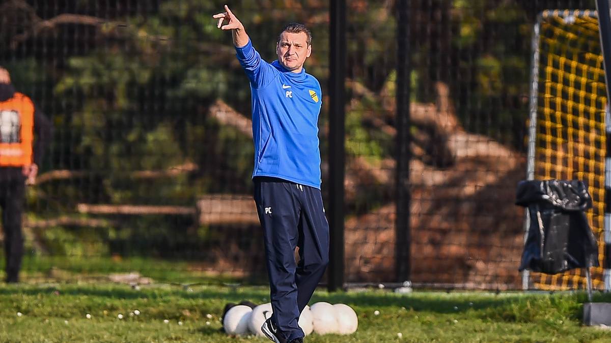 Trener w niebieskiej bluzie i granatowych spodniach stoi na boisku, wskazując ręką w górę. W tle widać siatkę bramki i inne osoby.