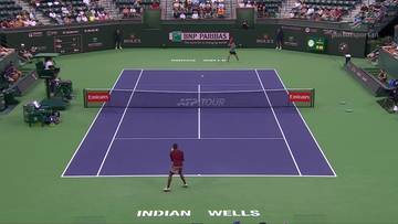 Kort tenisowy z zawodnikiem w czerwonym stroju przygotowującym się do uderzenia piłki. W tle widać trybuny z widzami i tablice informacyjne.