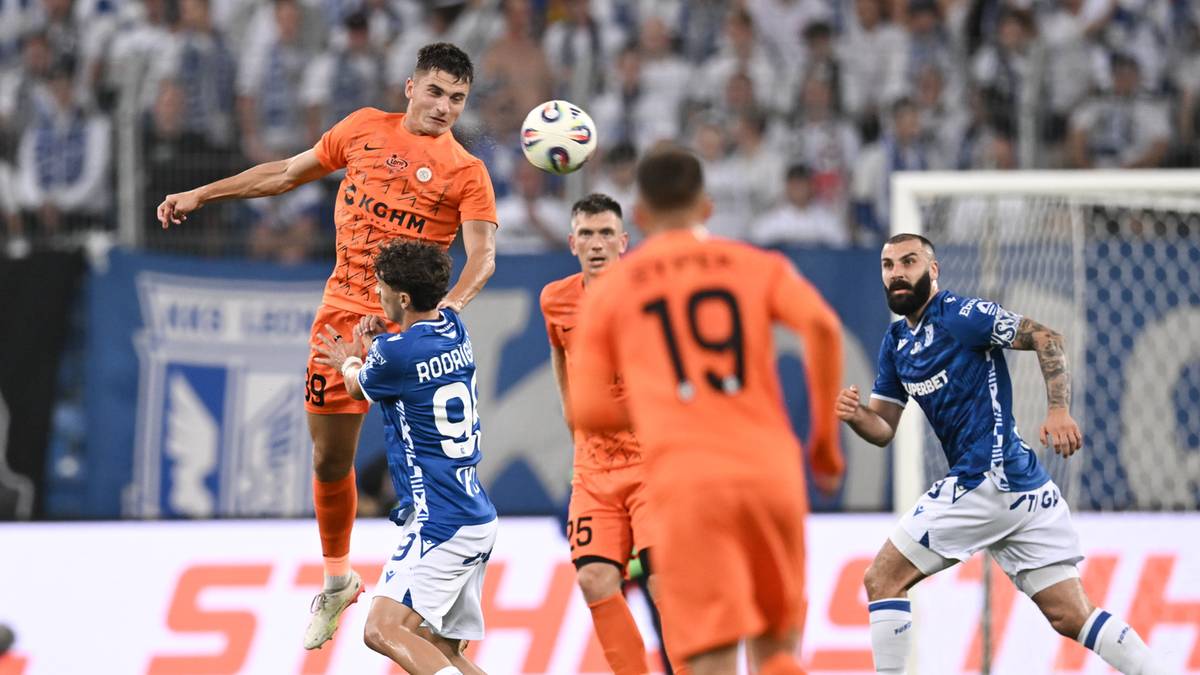 Gracz w pomarańczowym stroju wyskakuje do główki, celując w piłkę w powietrzu. Obok niego inni gracze w niebieskich i pomarańczowych strojach. Tło stanowi trybuna wypełniona kibicami.