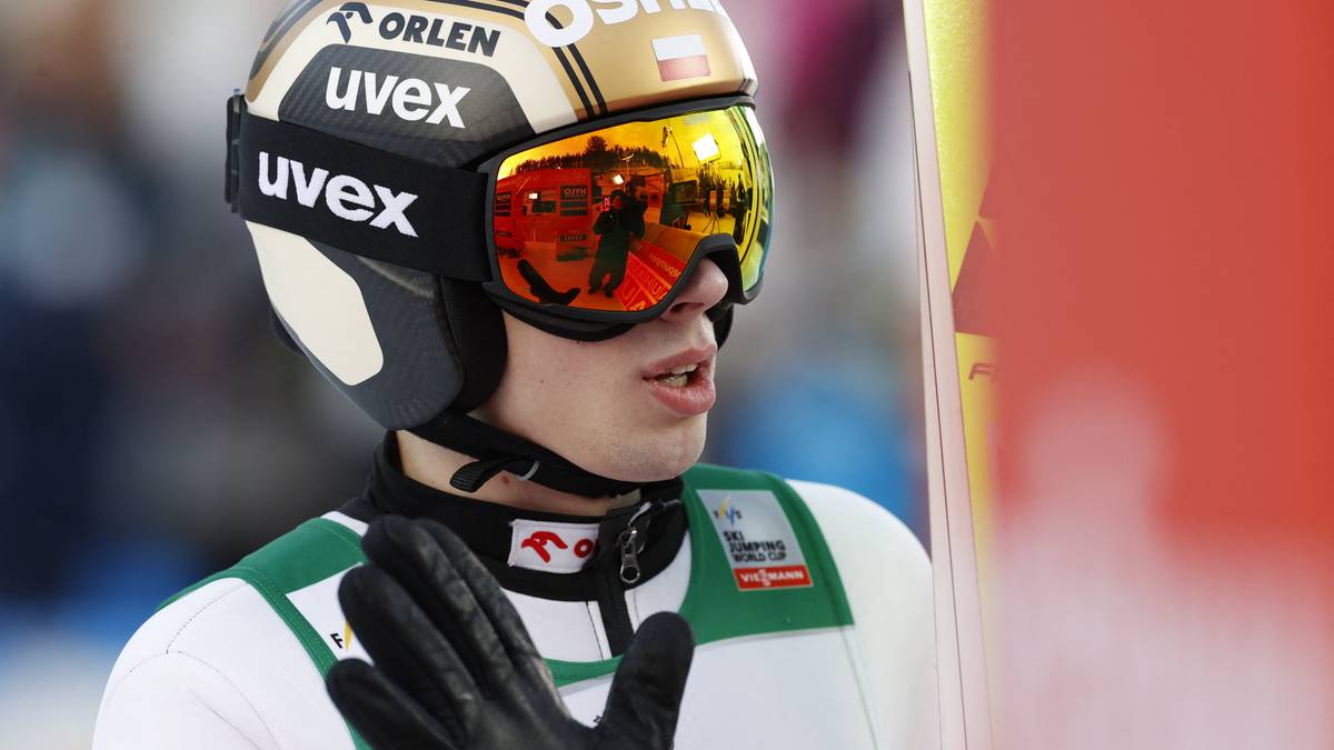 Skoczek narciarski w kasku i goglach z odbiciem publiczności w soczewkach, z rękawiczką na dłoni.