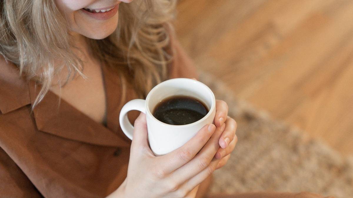 Kobieta trzymająca biały kubek z kawą w dłoniach, uśmiecha się do przodu.