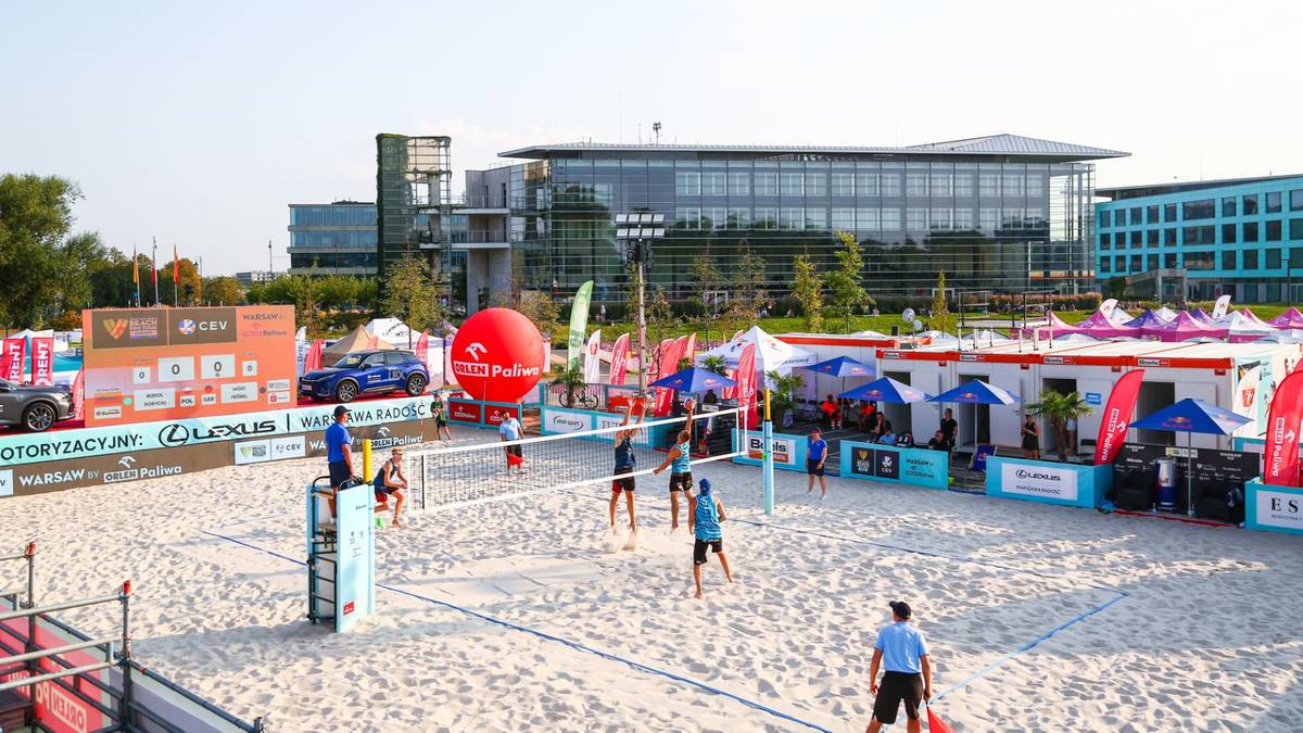 Boisko do siatk&oacute;wki plażowej z siatką, zawodnicy w akcji, czerwona piłka unosząca się nad boiskiem, widoczne trybuny i budynki w tle.