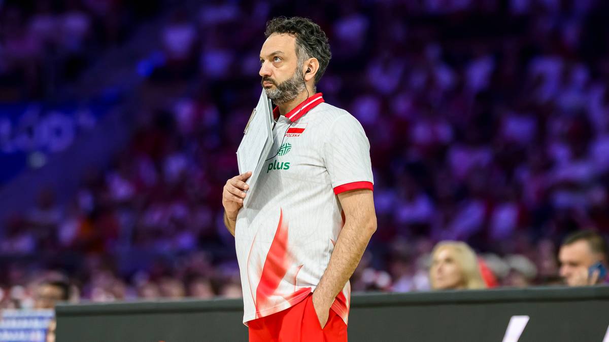 Trener w stroju sportowym z ręcznikiem w dłoni na tle trybun i boiska.