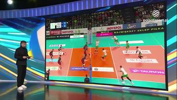 Duży ekran prezentujący mecz siatk&oacute;wki z graczami w akcji, boiskiem i trybunami pełnymi kibic&oacute;w. Obok ekranu stoi mężczyzna w czarnym garniturze.