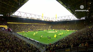 Stadion piłkarski wypełniony kibicami podczas meczu. Na murawie znajdują się piłkarze i ż&oacute;łte flagi.