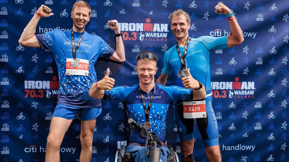 Trzech mężczyzn w strojach sportowych z medalami na szyjach uśmiecha się i pokazuje gesty radości. Dw&oacute;ch stoi, jeden siedzi na w&oacute;zku inwalidzkim. Tło stanowi baner zawod&oacute;w Ironman.