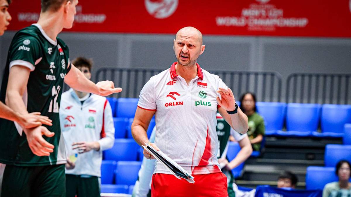 Trener w czerwono-białej koszulce Polskiego Związku Siatkówki z białym napisem "ORLEN PLUS" instruuje zawodników w zielonych strojach podczas meczu siatkówki.