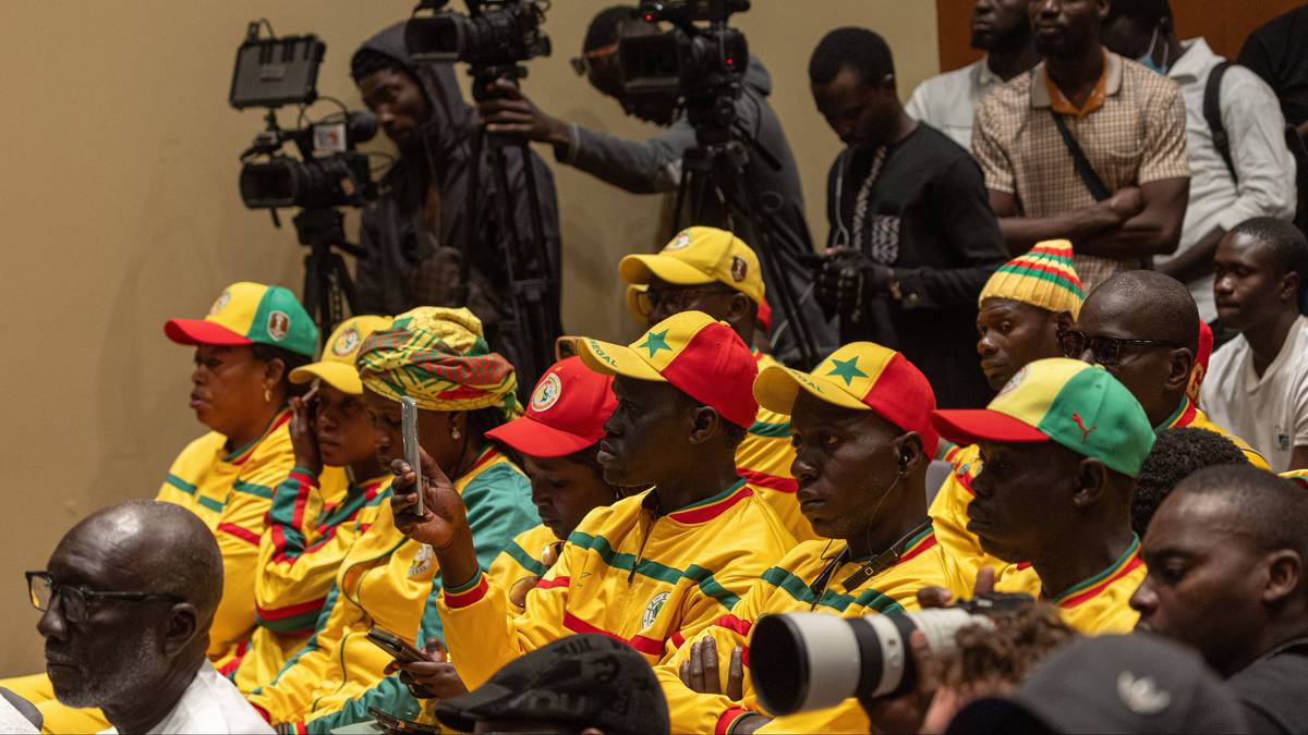 Grupa os&oacute;b w ż&oacute;łto-zielonych strojach z charakterystycznymi nakryciami głowy, z uwagą obserwująca coś poza kadrem. W tle widać osoby z kamerami.