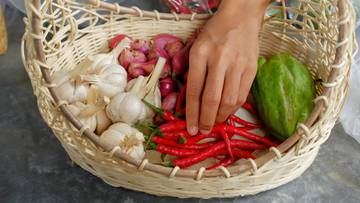 Koszyk z czosnkiem, szalotką, czerwonymi papryczkami i zielonym warzywem, do którego sięga ręka.