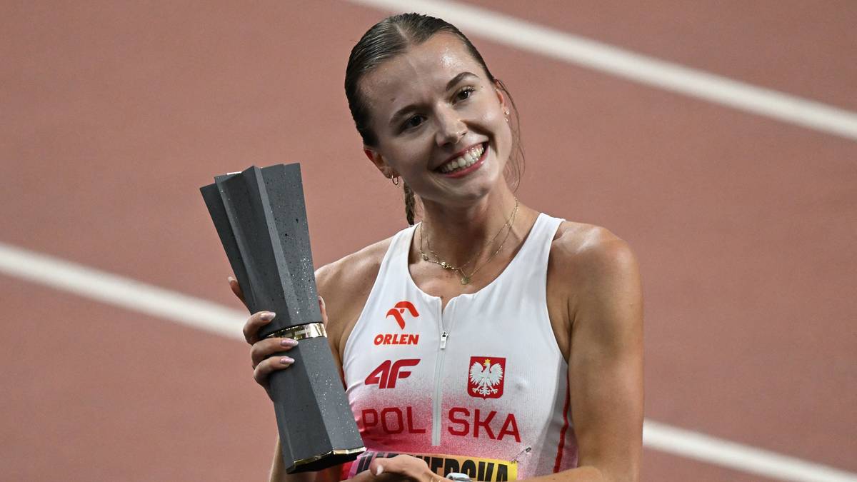 Sportsmenka w białej koszulce z napisem "POLSKA" trzyma w rękach szare trofeum i uśmiecha się.