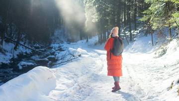 Kobieta w pomarańczowym płaszczu i czapce idzie zasypanym śniegiem leśnym szlakiem, obok płynie strumyk.