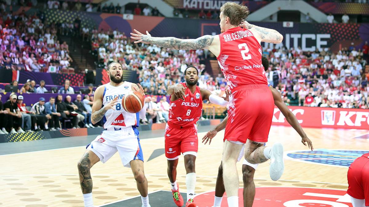 Trzech koszykarzy w akcji podczas meczu, jeden z piłką, dwaj pozostali próbują go zatrzymać. W tle trybuny wypełnione kibicami.