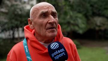 Starszy mężczyzna z łysą głową i zmarszczkami na twarzy, ubrany w pomarańczową bluzę, trzyma mikrofon Polsat Sport.