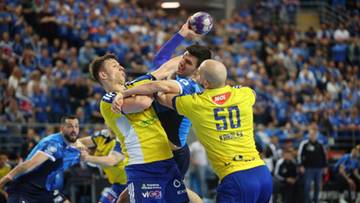 Trzech zawodników piłki ręcznej walczących o piłkę podczas meczu. Dwóch zawodników w żółtych koszulkach i jeden w żółto-niebieskiej koszulce. W tle trybuny zapełnione widzami.