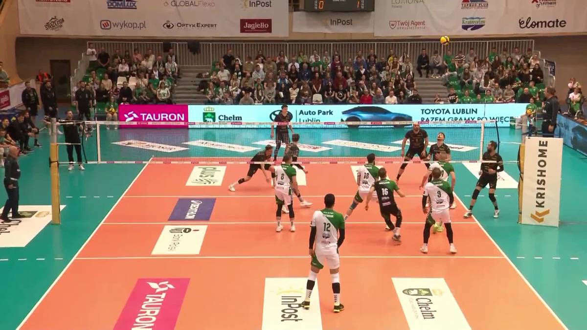 Mecz siatkówki z udziałem dwóch drużyn na boisku w hali sportowej. Zawodnicy w zielonych i białych strojach są w akcji, podczas gdy widzowie siedzą na trybunach w tle.