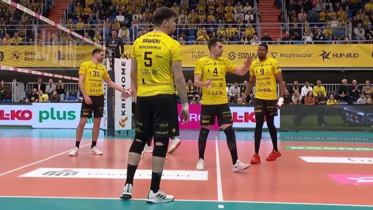 Czterech siatkarzy w ż&oacute;łtych koszulkach na boisku do siatk&oacute;wki. Dw&oacute;ch zawodnik&oacute;w stoi przodem do widza, jeden z nich ma numer 5 na plecach. Dw&oacute;ch innych zawodnik&oacute;w stoi bokiem, jeden z nich wykonuje gest ręką. Na drugim planie widać trybuny wypełnione widzami.