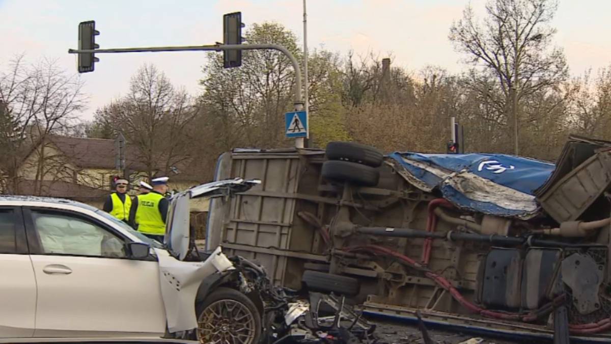 Wypadek drogowy ze skutkiem przewr&oacute;cenia się autobusu na bok, obok uszkodzony samoch&oacute;d osobowy i stojący policjanci.