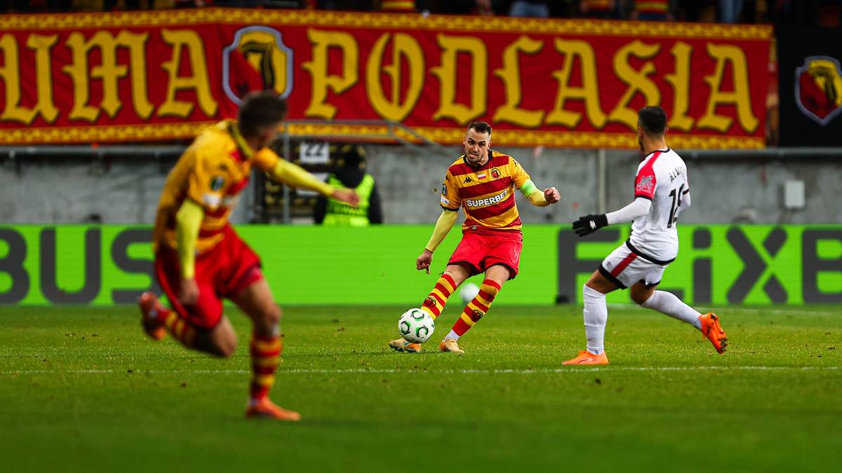 Trzech piłkarzy na zielonej murawie walczy o piłkę. Dw&oacute;ch zawodnik&oacute;w w czerwonych strojach i jeden w białym.