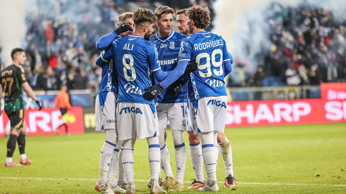 Piłkarze Lecha Poznań w niebieskich strojach zebrani w k&oacute;łku na boisku piłkarskim.