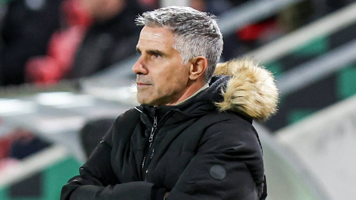 Trener piłkarski w czarnej kurtce ze złożonymi rękami na tle trybun stadionu.