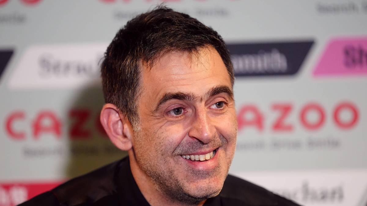 Portret Ronnie O'Sullivana, uśmiechniętego mężczyzny z ciemnymi włosami, w ciemnej koszuli, z logo "Cazoo" w tle.