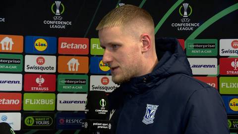 Portret mężczyzny o jasnych włosach i kr&oacute;tkiej brodzie, ubranego w granatową kurtkę z herbem klubu piłkarskiego. W tle znajduje się ściana z kolorowymi logotypami sponsor&oacute;w.