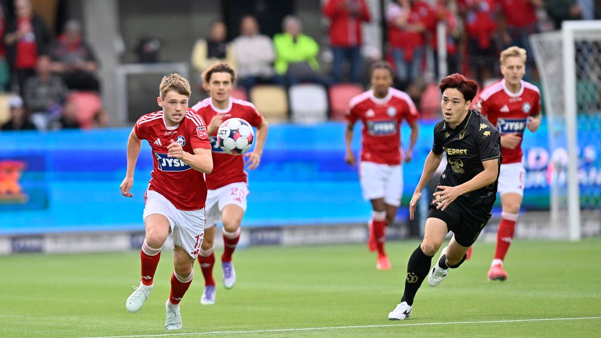 Piłkarz w czerwonej koszulce Jagiellonii Białystok biegnie z piłką na boisku piłkarskim, ścigany przez dwóch zawodników w czarnych koszulkach Silkeborg IF. W tle widać innych graczy oraz trybuny stadionu.