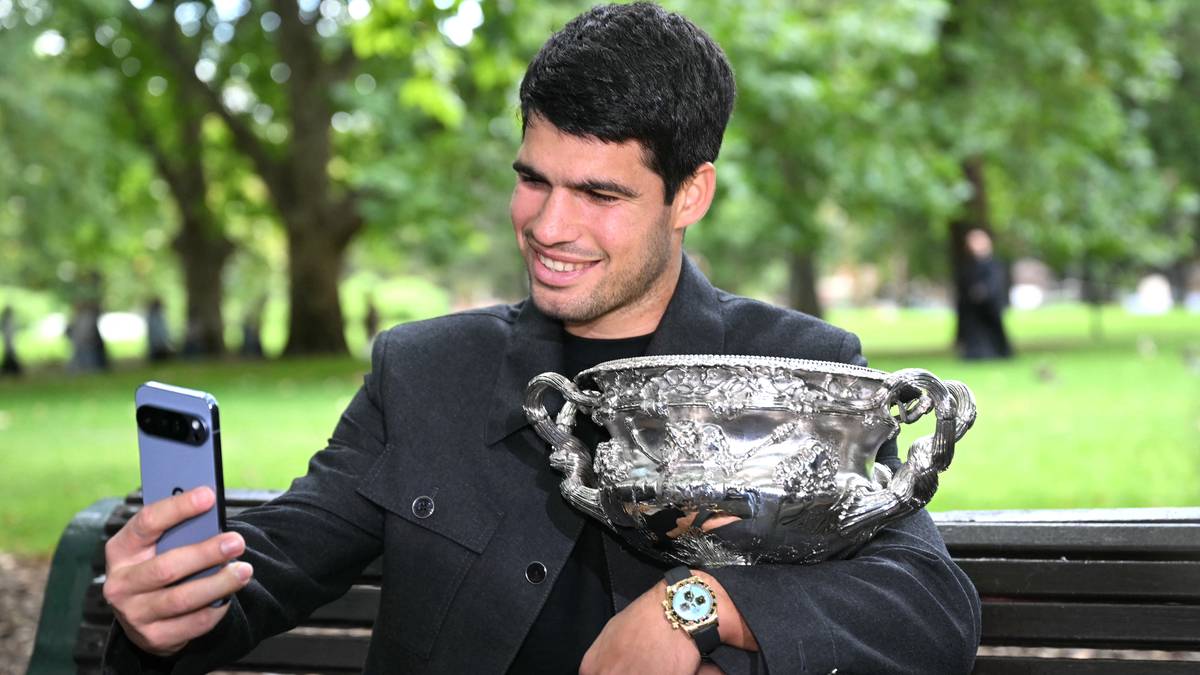 Młody mężczyzna w garniturze siedzi na ławce, trzymając w jednej ręce trofeum, a w drugiej smartfon, robiąc sobie zdjęcie.