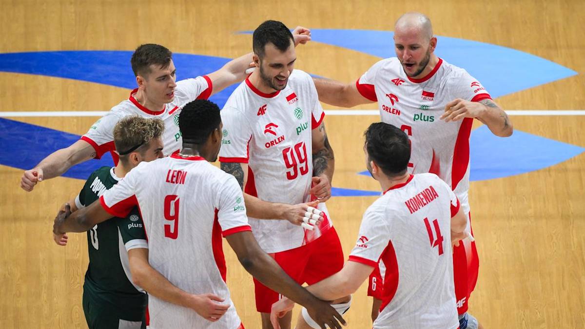 Siedmiu mężczyzn w strojach sportowych, w większości w kolorze białym z czerwonymi elementami, świętuje na drewnianej hali sportowej.