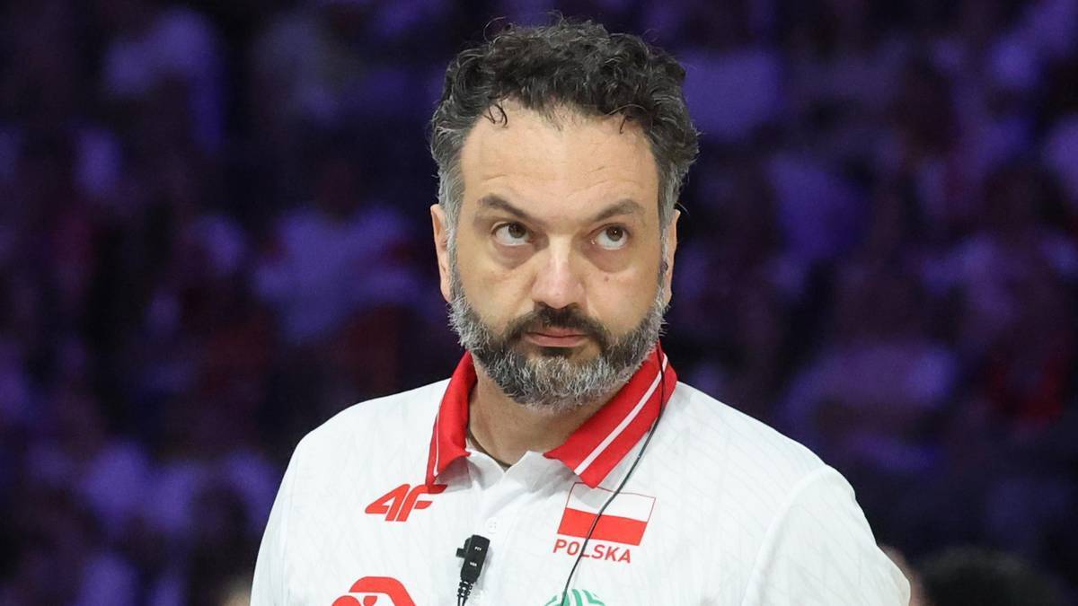 Mężczyzna z brodą i kręconymi włosami, ubrany w białą koszulkę sportową z czerwonym kołnierzykiem i godłem Polski na piersi, patrzy w bok z poważnym wyrazem twarzy.