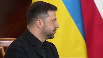 Wołodymyr Zełenski, prezydent Ukrainy, w czarnym garniturze, siedzi na tle flagi Ukrainy i Polski.