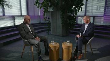 Dw&oacute;ch mężczyzn w garniturach siedzi naprzeciwko siebie na krzesłach w pomieszczeniu z przeszklonymi ścianami.