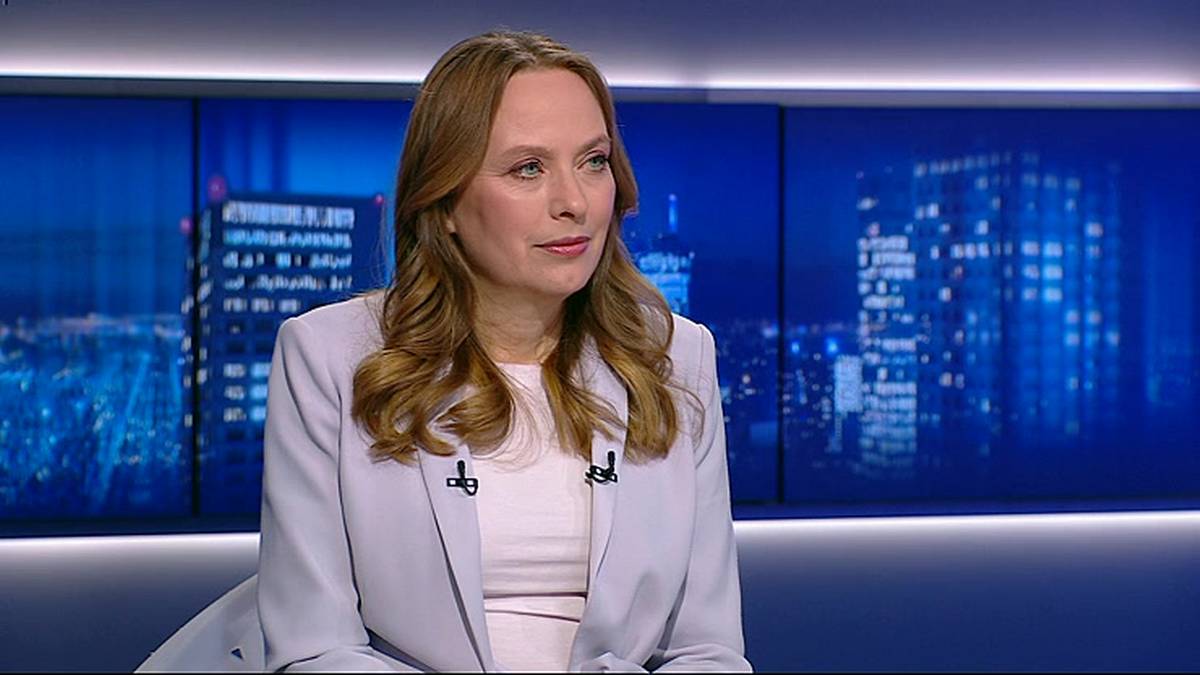 Kobieta w jasnej marynarce i białej bluzce siedzi, patrząc w prawo. Tło stanowią niebieskie ekrany z widokiem na nocne miasto.